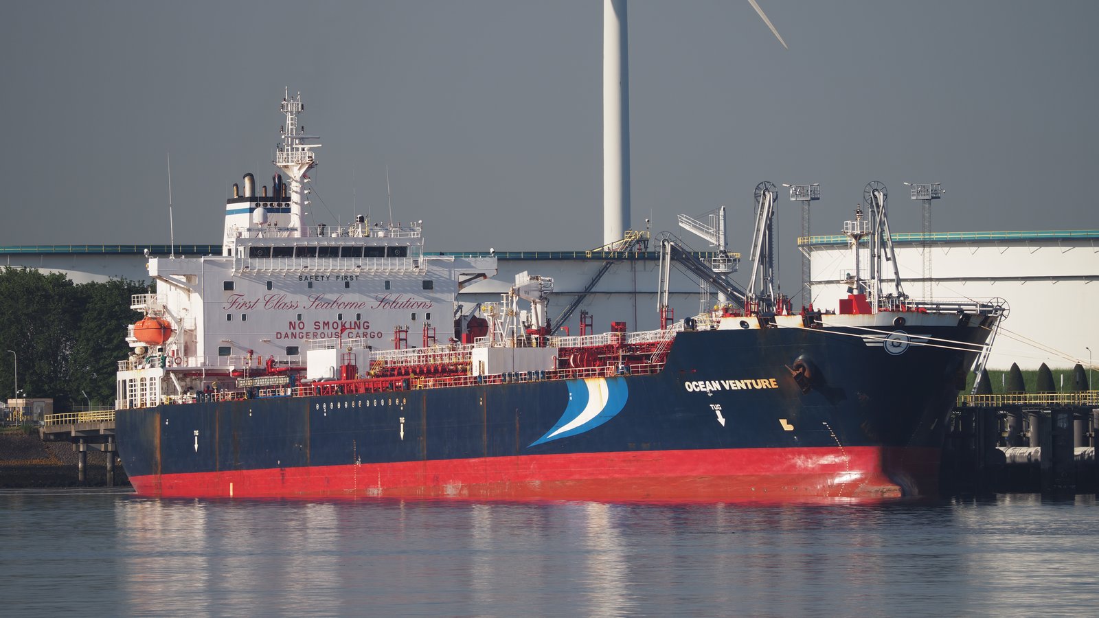 A dark blue and red oil and chemical tanker moored at a quay beside white storage tanks, with a wind turbine behind it